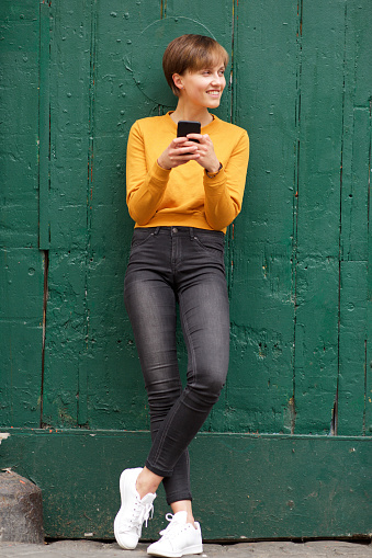 Full body portrait of young woman  leaning against wall with mobile phone