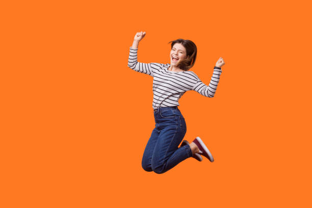full length portrait of excited pretty woman with brown hair in casual shirt and denim jumping. indoor studio shot isolated on orange background - women female cheerful ecstatic imagens e fotografias de stock