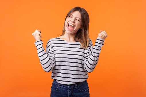 Yes I did it! Portrait of joyous winner, young woman with brown hair in casual shirt standing with clenched fists and closed eyes, celebrating victory. indoor studio shot isolated on orange background