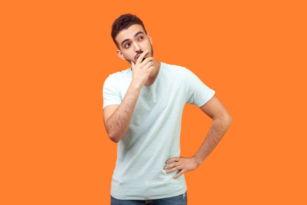 Need to think. Portrait of thoughtful brunette man holding his chin and pondering idea. indoor studio shot isolated on orange background Need to think. Portrait of thoughtful brunette man with beard in white t-shirt holding his chin and pondering idea, confused not sure about solution. indoor studio shot isolated on orange background grimacing photos stock pictures, royalty-free photos & images