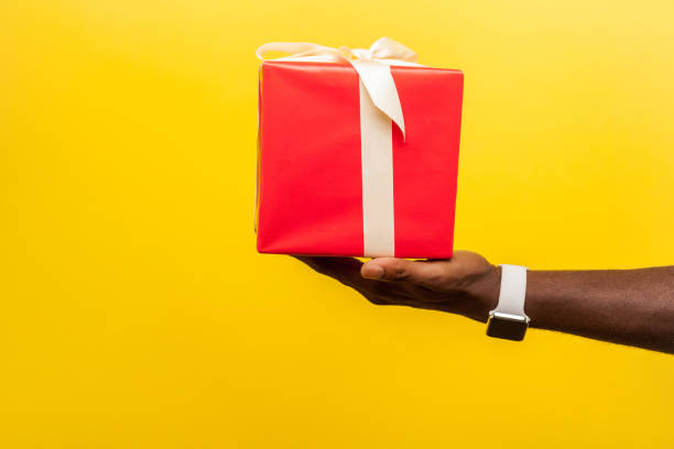 Closeup side view of male hand with wristwatches holding out red gift box. isolated on yellow background Closeup side view of male hand with wristwatches holding out red gift box with ribbon, giving present on holiday, bonuses and surprises concept. indoor studio shot isolated on yellow background hand holding gift stock pictures, royalty-free photos & images