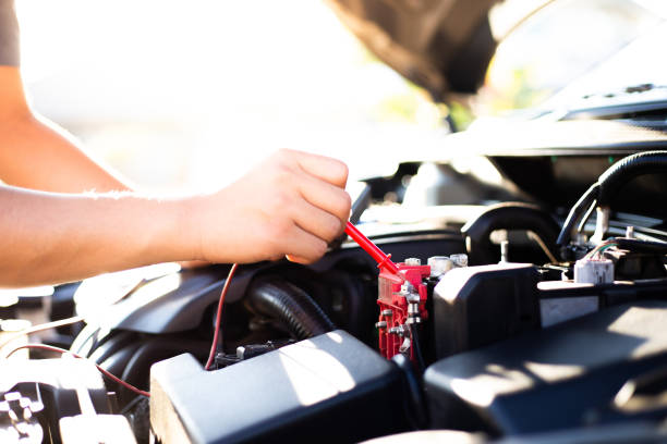 54 year old man in glasses wearing red and black winter jacket examines car engine with open hood in forest residential setting. Red car with open hood stands in wooded backyard during winter season. House visible in background through pine trees