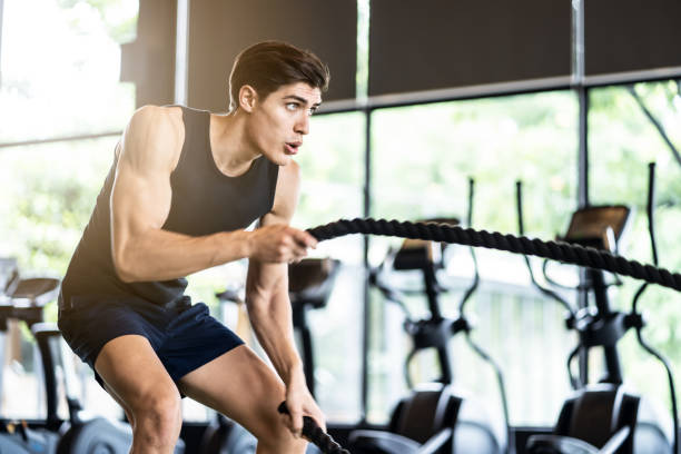 Young fit athletic sporty Caucasian man in sportswear working out in Gym or fitness club. The man swinging battle ropes firmly showing his power and strength expression on face. Seen from model side. joven ajuste deportivo deportivo hombre caucásico en ropa deportiva haciendo ejercicio en gimnasio o gimnasio. el hombre balanceando cuerdas de batalla mostrando firmemente su poder y expresión de fuerza en la cara. visto desde el lado del modelo.