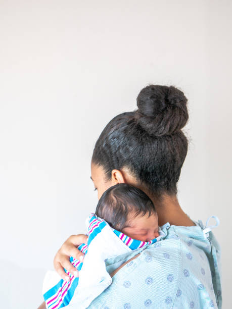 A mixed race African American mother faces away from the camera wearing a hospital gown holds her brand new infant baby over her shoulder hugging and cradling him as he sleeps in his blanket swaddle. stock photo