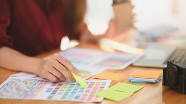 istock Cropped side shot of young graphic designer while selecting color code from the color guide on the work desk. Including camera, paper note, laptop and color guide on work desk. 1198158969