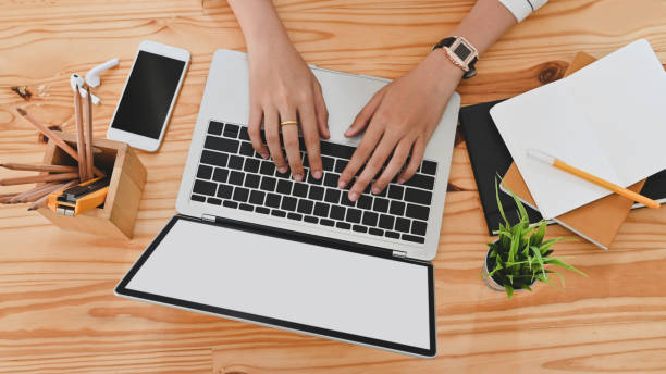 istock Top view of woman hand while using/typing on black blank screen laptop including pencil holder, black blank screen smartphone, potted plant, pencil, notes, earphone on wooden desk. 1198159003