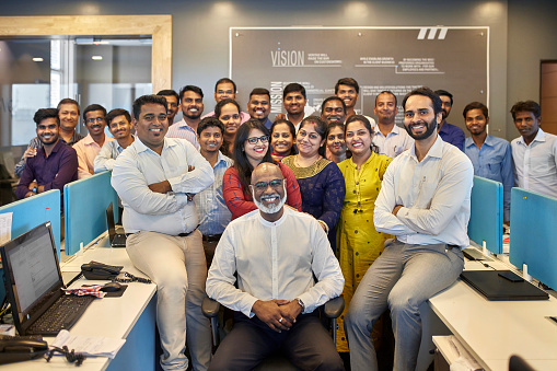 Senior Indian businessman sitting in chair surrounded by smiling senior managers with arms crossed and standing staff workers.