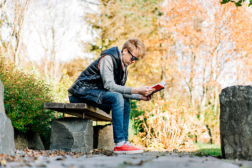 Full length of teenage boy reading novel while sitting on bench. Side view of fashionable male teenager is at park. He is wearing warm clothing.