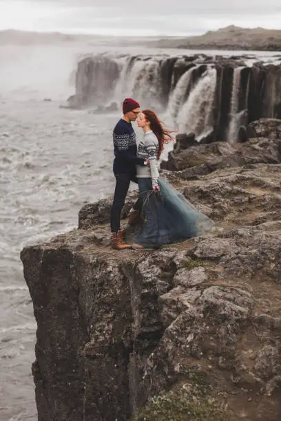 Photo of Happy hipster couple in love walking near popular Selfoss waterfall in Iceland. Traditional wool sweaters, red hair, gray skirt. Dramatic nordic landscape, cold weather.