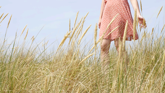 Body shot of a young woman wearing a summer dress walking through the long grass by the sand dunes on a beach.