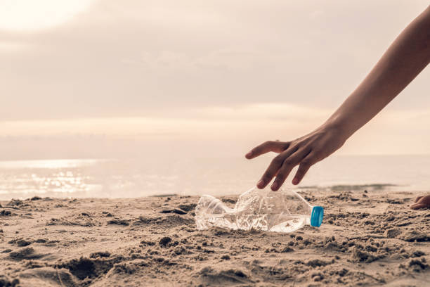 istock Cropped Hand Of Man Picking Plastic Bottle At Beach Against Sky 1198210695