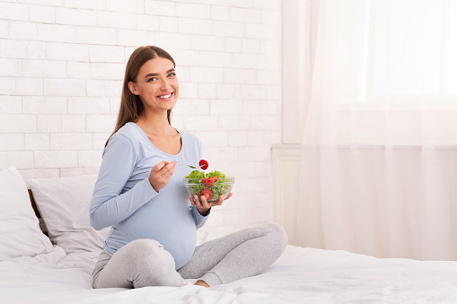 Pregnancy Healthy Diet. Pregnant Girl Holding Vegetable Salad In Bowl Smiling To Camera Sitting In Bed At Home. Empty Space