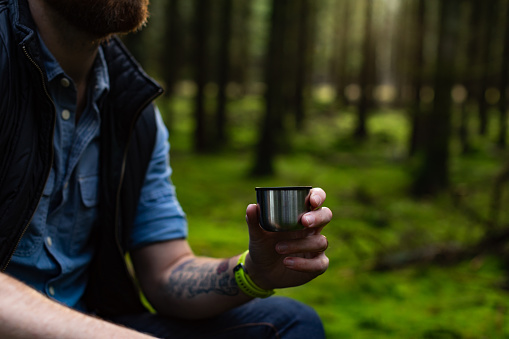A caucasian man is drinking coffee in a green forest in Sweden during springtime. The picture is a close-up of the man holding a silver mug.