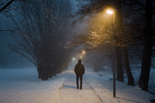 Young man alone walking on sidewalk under yellow street lights in mist. Foggy air. Peaceful atmosphere in snowy night. Back view.