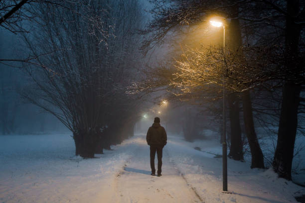молодой человек один ходит по тротуару под желтыми уличными огнями в тумане. туманный воздух. мирная атмосфера в снежную ночь. вид сзади. - park snow tree back lit стоковые фото и изображения