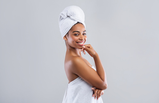 Cheerful young african woman wrapped in white bath towels after shower, grey studio background, free space