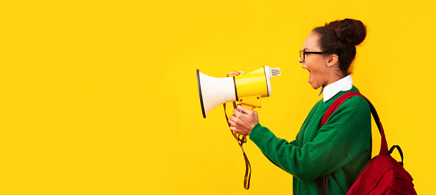 Attention. Black teen girl with megaphone screaming isolated over yellow background, panorama