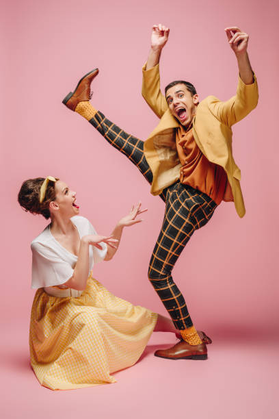 cheerful girl sitting on floor and excited man raising leg while dancing boogie-woogie on pink background cheerful girl sitting on floor and excited man raising leg while dancing boogie-woogie on pink background boogie woogie dancing stock pictures, royalty-free photos & images