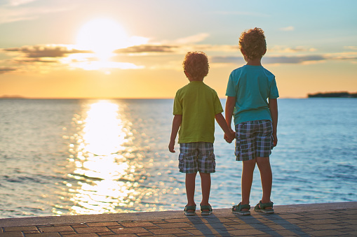 Two cute red curly boys brothers standing on the seashore looking at beautiful sunset sky.