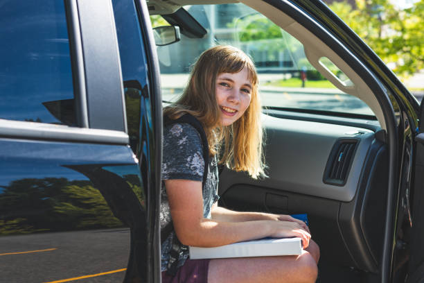 Happy teenage girl sitting/stepping out of a parked car/suv while getting dropped off/picked up from school. stock photo