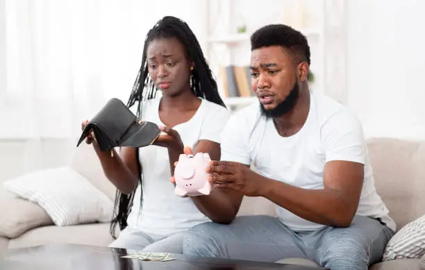 Photo of Unhappy afro couple shaking piggybank and wallet at home, searching money