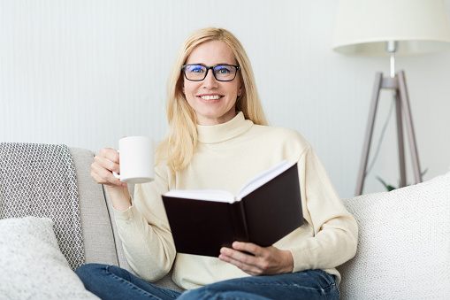 Excited middle-aged woman drinking tea and reading book on sofa, smiling to camera