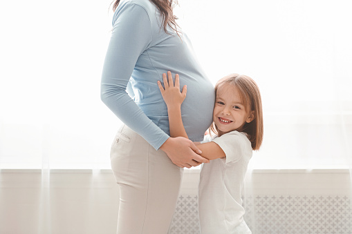 Close up of little daughter embracing her pregnant mom, cute girl touching expecting mother tummy