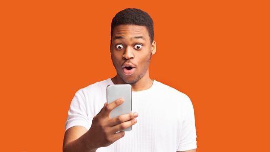 Shocking News. Anxious black man looking at phone seeing bad news or photos over orange background, panorama