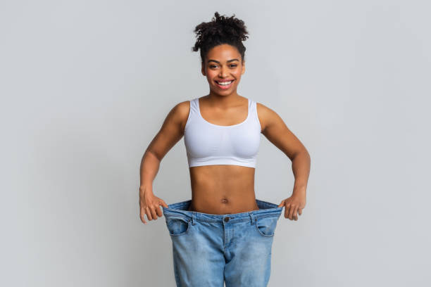 Happy young woman with too large jeans Successful weight loss, afro woman with too large jeans after effective diet, grey background too thin stock pictures, royalty-free photos & images