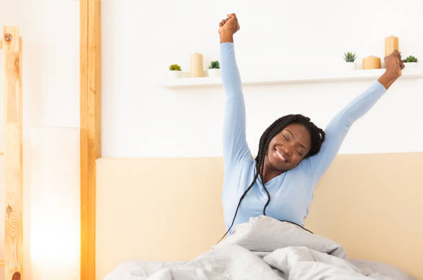 senhora feliz que estica os braços que acordam na cama em casa - wake up stretching women black - fotografias e filmes do acervo