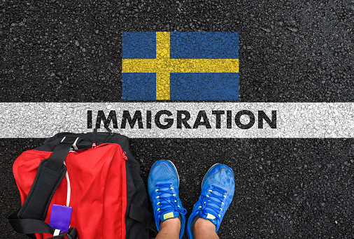 Man in shoes with bag standing next to line with word IMMIGRATION and flag of Sweden on asphalt road