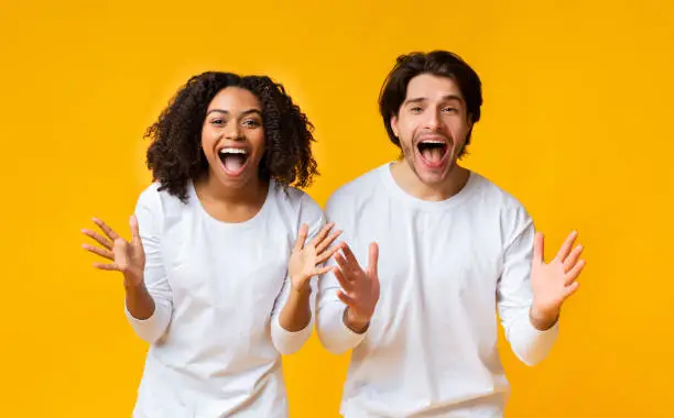 Shocked Multiracial Couple Exclaiming With Amazement And Emotionally Gesturing With Hands Over Yellow Background With Empty Space