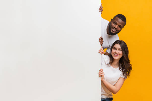 istock Joyful interracial couple peeking from behind white advertisement board 1198237443
