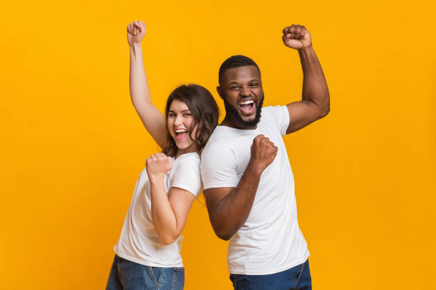 Joyful interracial couple rejoicing success, raising fists, celebrating victory We are the champions. Joyful interracial couple rejoicing success, raising fists, celebrating victory, posing together over yellow background clingy girlfriend stock pictures, royalty-free photos & images