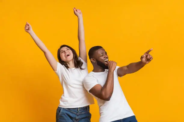 Joyful international couple having fun together, emotionally dancing and singing over yellow background with empty space