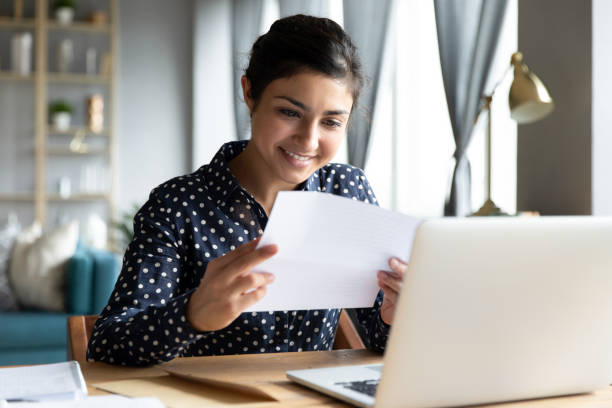 Smiling indian woman holding reading paper letter sit at table Smiling young indian woman holding paper bill letter doing paperwork bill reading good news check post mail sit at home table, happy lady customer receive bank receipt sheet tax refund notification financial statement stock pictures, royalty-free photos & images