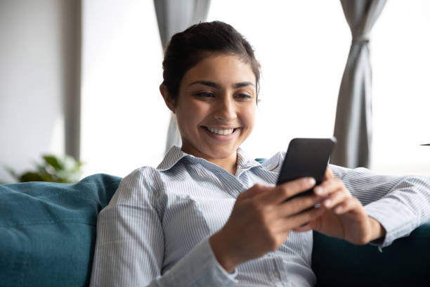 istock Smiling indian girl using smart phone mobile apps on sofa 1198252620