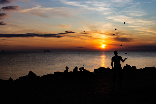 Juggler performance with many balls on the beach of the mediterranean sea in trieste, italy while sunset. Only silhouette visible at dusk. Couple and other people sitting at the coast line.