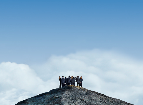 A rear view of a group of business people as they look in different directions while standing at the peak of a mountain top.