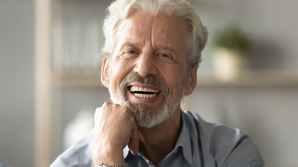 Portrait elderly man with candid wide smile looking at camera Close up portrait handsome face of 60s elderly man having candid wide toothy smile put fist under chin looking at camera concept of healthy person enjoy retired life, dentures services for old people dentures stock pictures, royalty-free photos & images