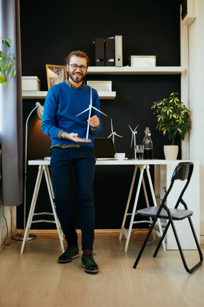 istock Young handsome positive smiling caucasian engineer standing in his office, showing windmill model and looking at camera. 1198267101