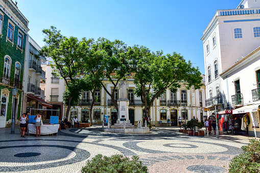 Lagos, Portugal - 07.18.2019: Houses in the old town, square tiled with calçada portuguesa with patterns in the form of black circles, green trees, people walk, in the summer afternoon.