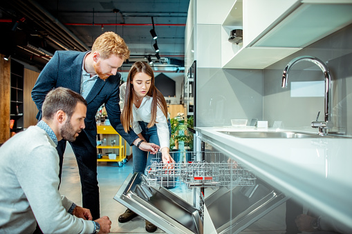 Shop salesman explaining the features of a new dishwasher model to a young shopping couple in a kitchen appliances store.