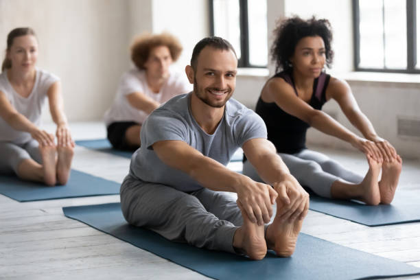 Smiling man practicing yoga at group lesson, Seated forward bend Smiling young man instructor wearing grey sportswear practicing yoga at group lesson, doing Seated forward bend exercise, diverse people stretching in paschimottanasana pose on mats, working out hamstring stock pictures, royalty-free photos & images