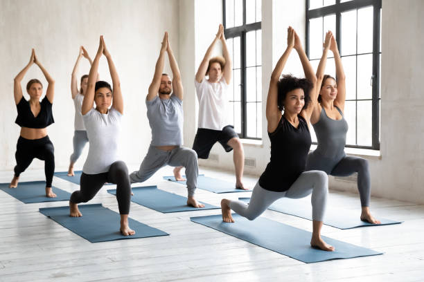 Young diverse people doing Warrior one exercise, practicing yoga Young diverse young people with African American instructor doing Warrior one exercise, practicing yoga at group lesson, standing in Virabhadrasana pose, working out in modern yoga studio yoga class stock pictures, royalty-free photos & images
