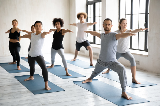 Young diverse people wearing sportswear doing Warrior two exercise at group lesson, practicing yoga, standing in Virabhadrasan pose on mats, working out with instructor in modern yoga studio
