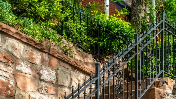 Pano frame Outdoor staircase with stone steps and black metal railing against a fence. Close up of a sunlit stairway with house and plants at the top.
