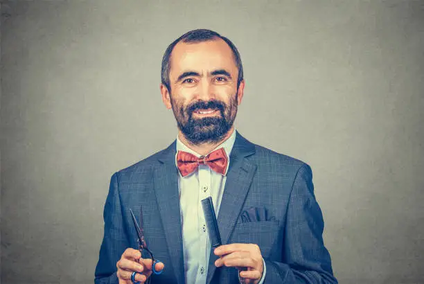 Smiling bearded hairdresser wearing elegant jacket with red bow tie, posing with scissors and comb. Stylish adult man studio portrait, isolated at gray background. Human face expression