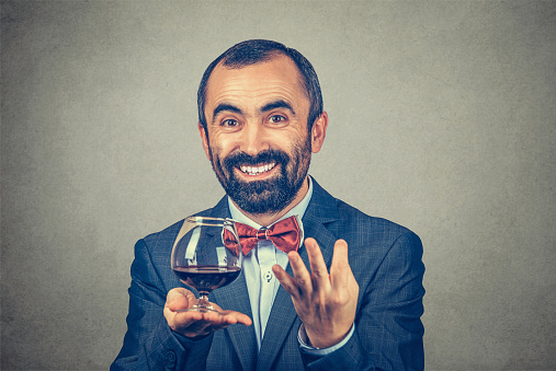 Smiling adult bearded italian man wearing elegant jacket with red bow tie with wine glass looking at camera asking what do you say with hand gesture isolated grey background. Positive human expression