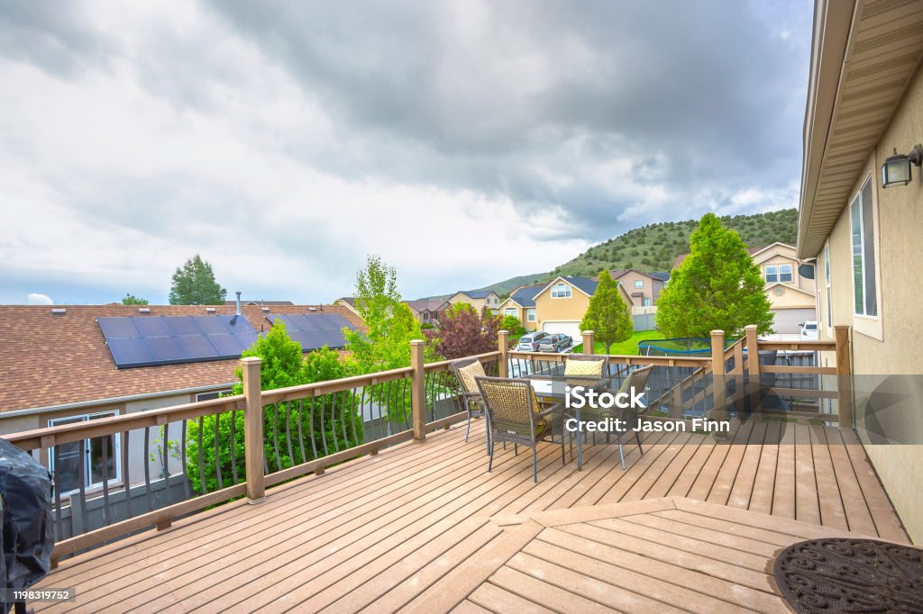 Deck overlooking solar panels on roof of home against mountain and cloudy sky Deck overlooking solar panels on roof of home against mountain and cloudy sky. The deck has wooden floor, furniture, and stairs that leads down to the yard. Deck Stock Photo